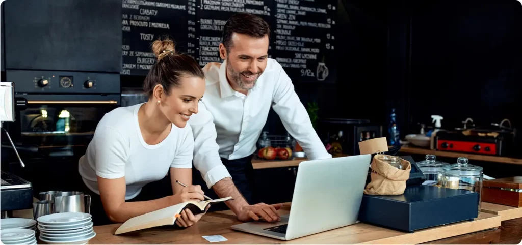 A man and woman work on their laptop at the restaurant they work at to monitor and respond to online reviews as part of their restaurant's online marketing strategy.